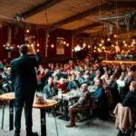 A speaker addressing a large crowd in a vibrant, warmly-lit indoor venue.