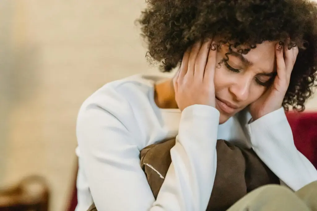 Frustrated African American female with closed eyes touching face while sitting with cushion on couch in room on blurred background