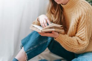 Crop unrecognizable female in warm sweater sitting on floor and reading interesting book in light room